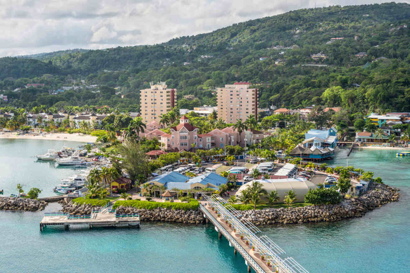 Aerial view of a coastal town with buildings, a pier, and boats docked in a harbor, surrounded by lush green hills and the sea.