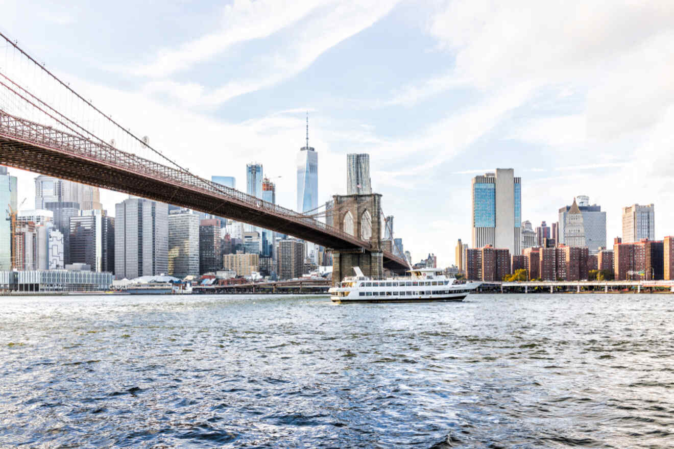 Brooklyn Bridge spans across the river with a ferry passing underneath. The city skyline, including tall buildings, is visible in the background under a partly cloudy sky.