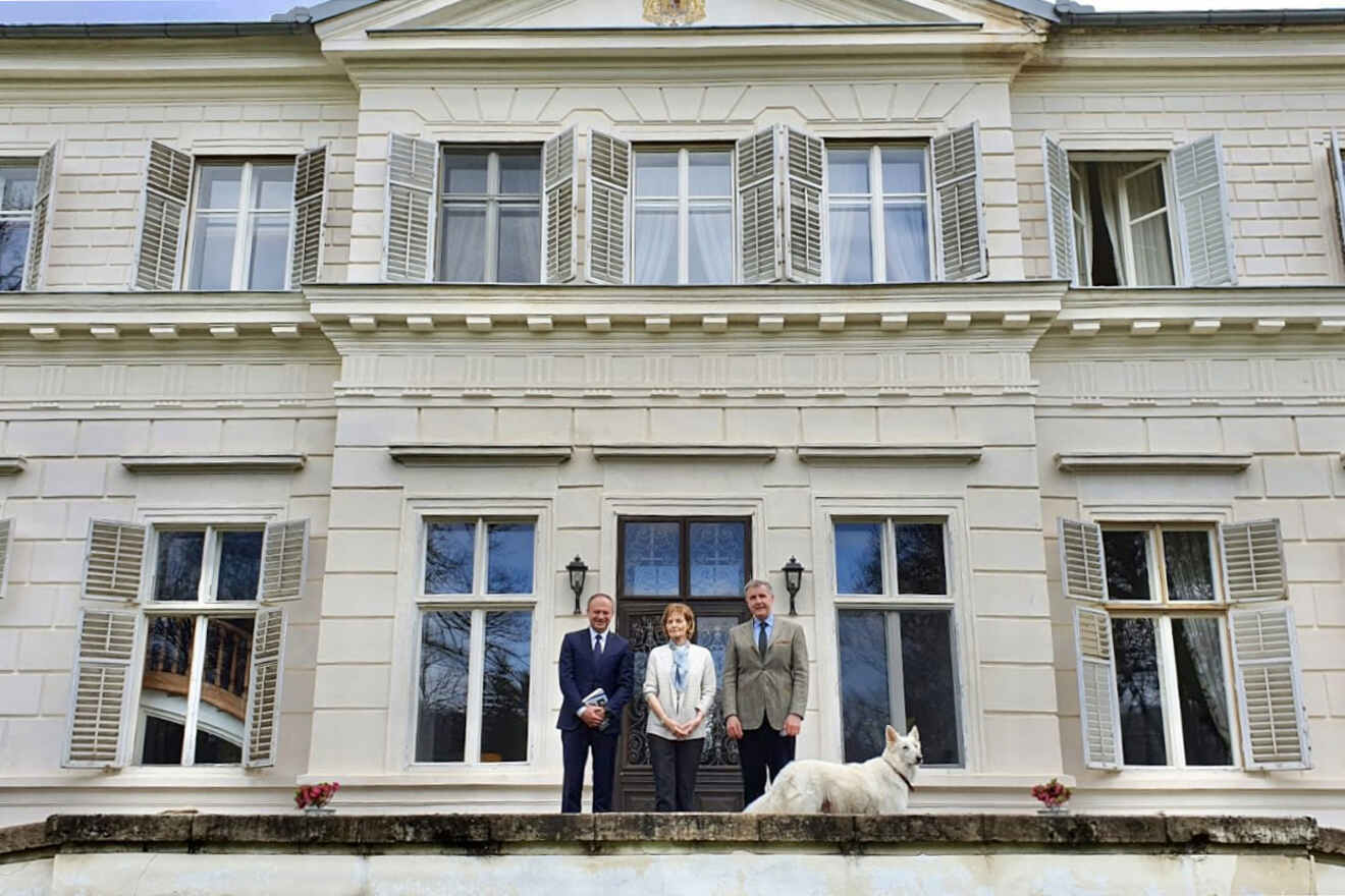 Four people and a dog stand in front of a large, white, two-story building with shuttered windows.