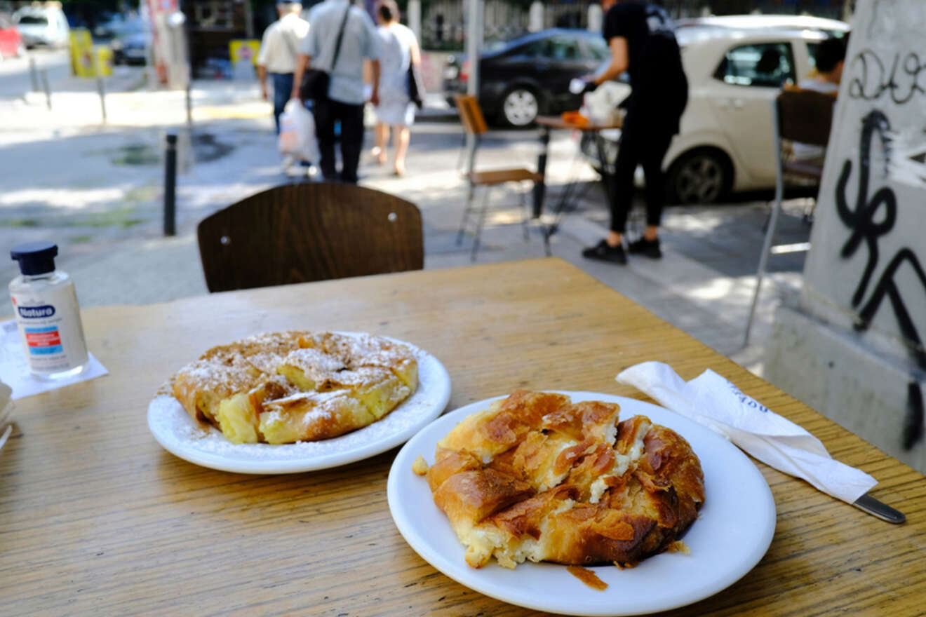 Two plates of pastry sit on a wooden table outdoors, with a bottle of hand sanitizer nearby. In the background, people walk on a sunny street.