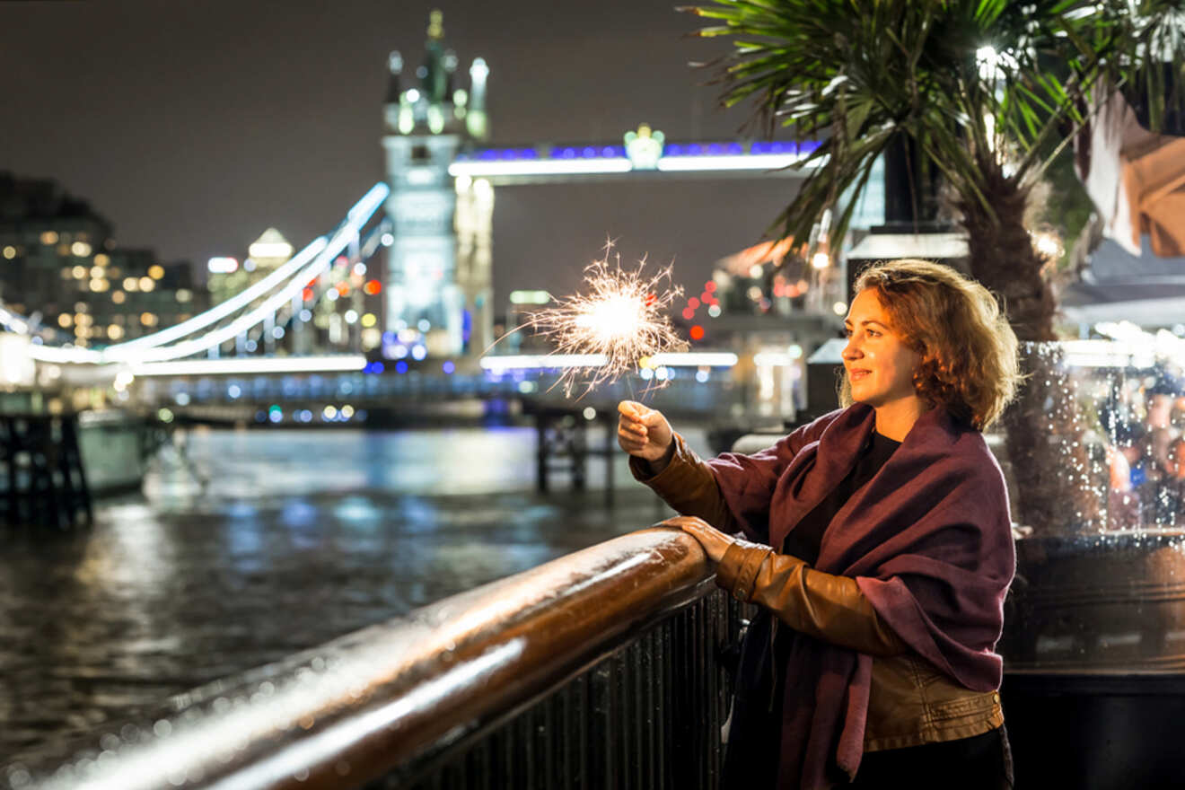 A woman holds a sparkler by the riverside at night, with an illuminated bridge in the background.