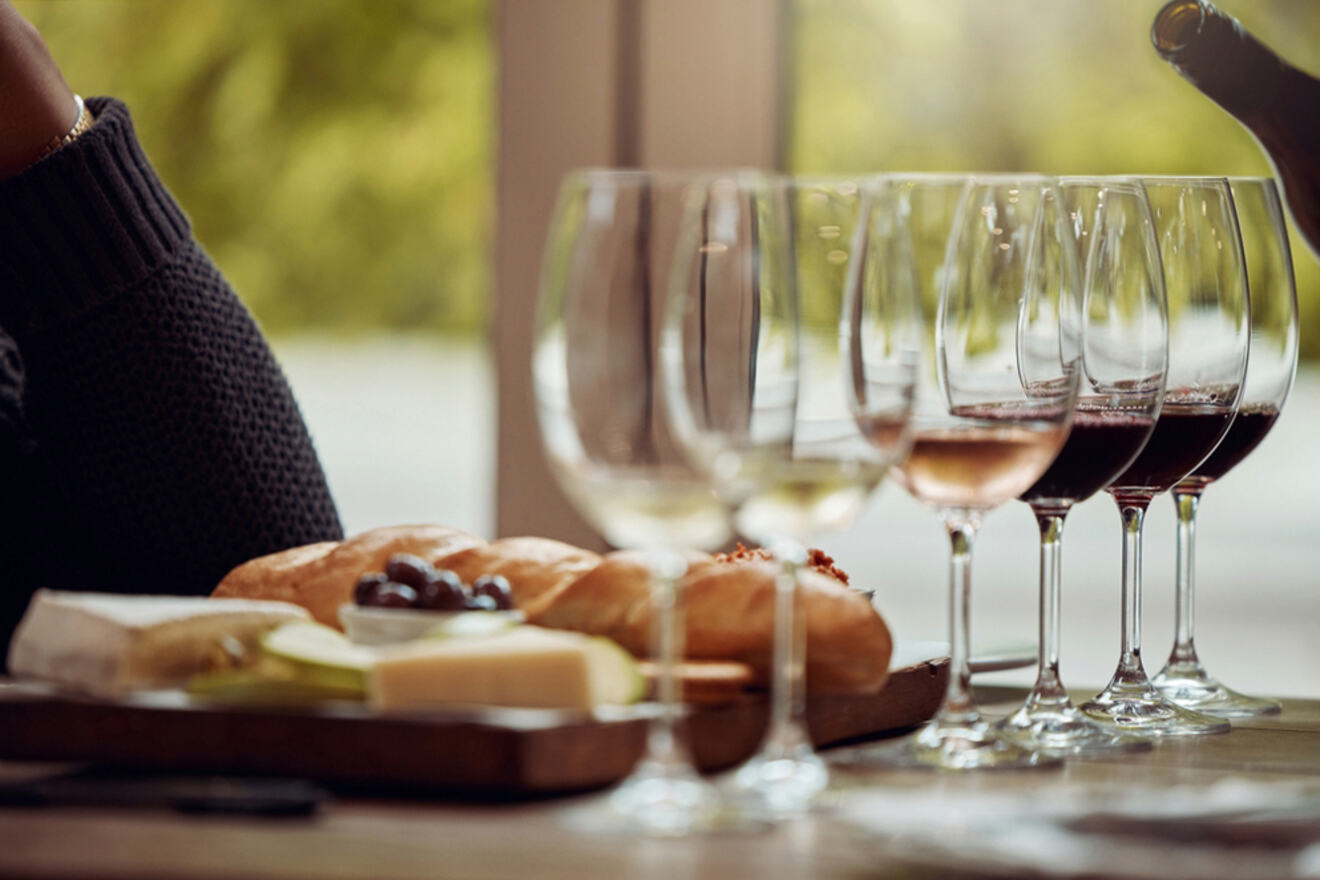 A row of wine glasses with red, white, and rosé wine sits in front of a platter of bread, cheese, and olives on a wooden table, reminiscent of a sunlit afternoon in Lagos, Portugal.