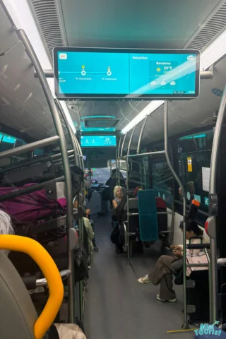 Inside a bus, passengers are seated. A screen displays weather information for Barcelona (22°C) and other locations. Luggage is stored on racks above seats.