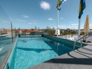 Rooftop swimming pool with glass sides overlooking a cityscape under a blue sky, with flags and buildings visible in the background.