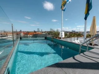 Rooftop swimming pool with glass sides overlooking a cityscape under a blue sky, with flags and buildings visible in the background.