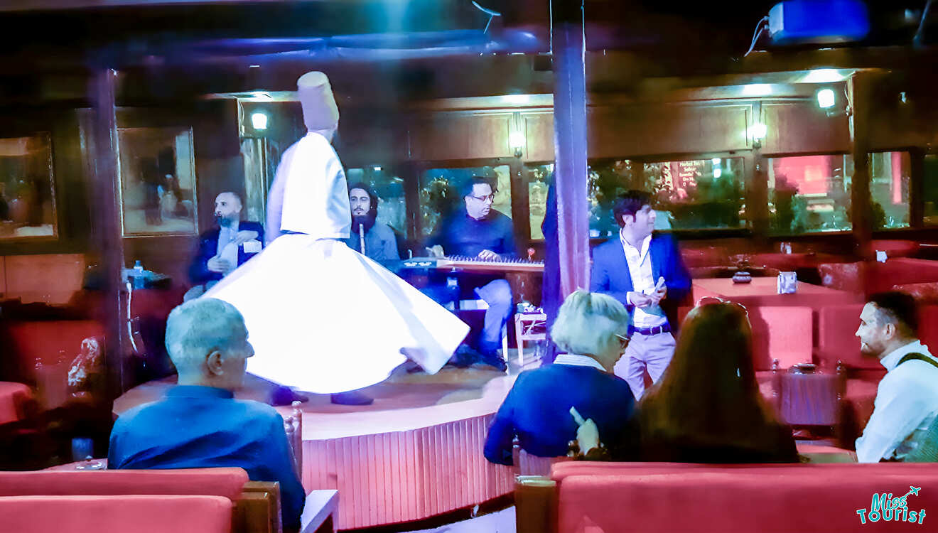 A dervish dancer spins on stage while musicians perform. An audience watches in a dimly lit venue with red seating.