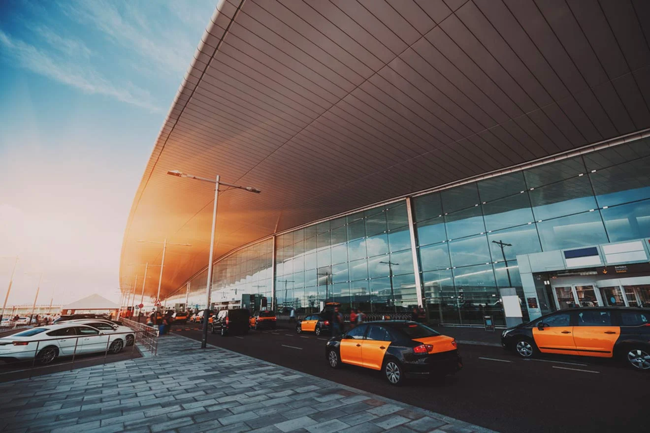 A row of taxis is parked outside a modern airport terminal with a curved roof and large glass windows.