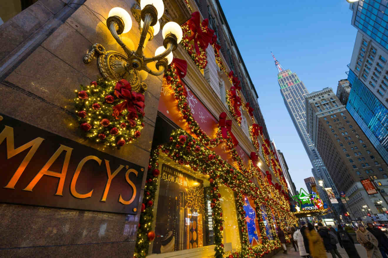 Macy's store decorated with festive lights and ribbons, with the Empire State Building visible in the background and people walking on the sidewalk.