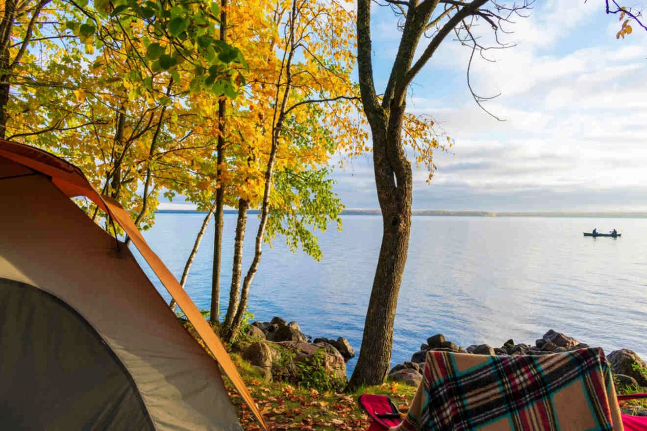 A campsite by a lake with a tent, a chair, and a plaid blanket. Trees with autumn foliage, rocks by the water, and a boat in the distance.