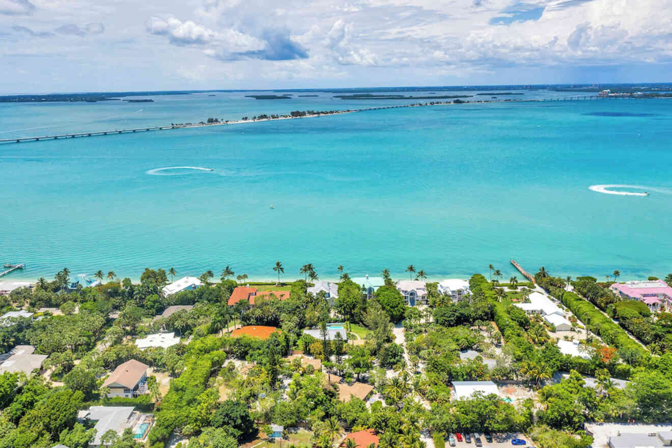 Aerial view of a coastal area with turquoise waters, a thin island strip, a bridge, and residential buildings surrounded by greenery.