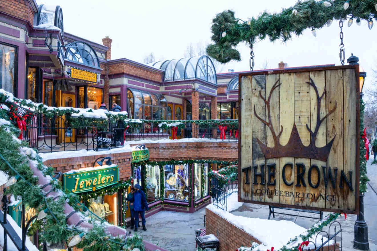 A snowy outdoor shopping area with shops like jewelers and a coffee bar. Festive decorations and a sign for "The Crown" are visible.