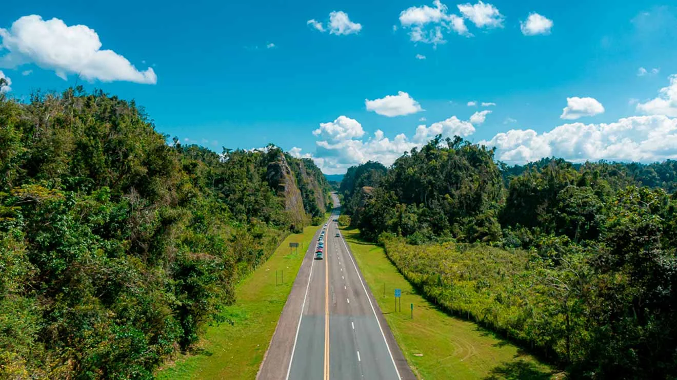 A highway cuts through lush green hills under a blue sky with scattered clouds. Cars travel along the road.