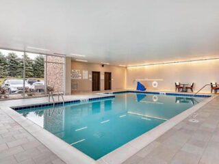 Indoor swimming pool with clear water, tiled deck, pool ladder, safety ring, and a few tables and chairs; large window shows parked cars and greenery outside.