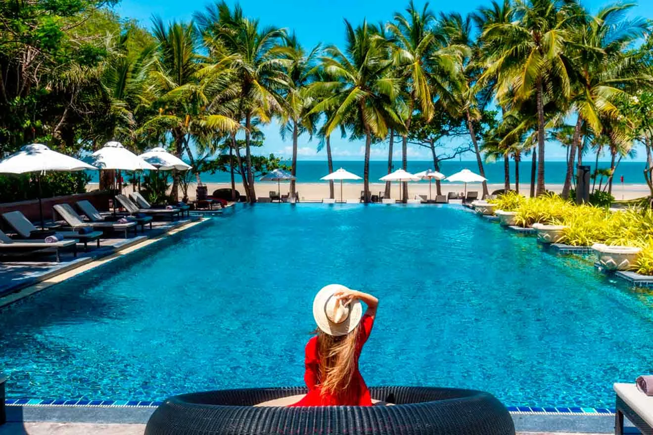 A woman in a red dress sits by a pool, facing the ocean. She is wearing a hat. The scene is surrounded by palm trees and beach umbrellas.