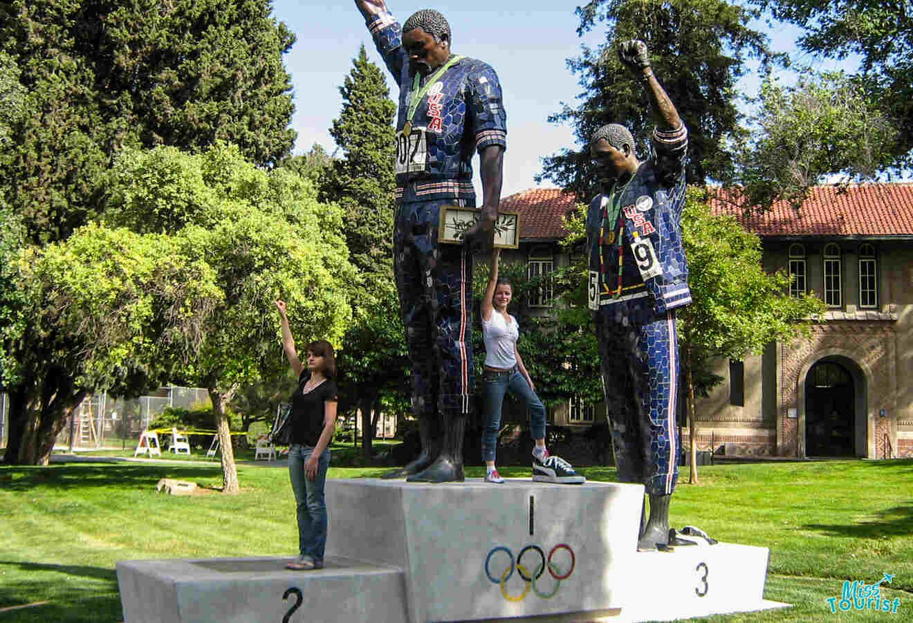 The writer of the post with her friend mimic a historic Olympic podium statue depicting athletes raising their fists, set outdoors on a sunny day.