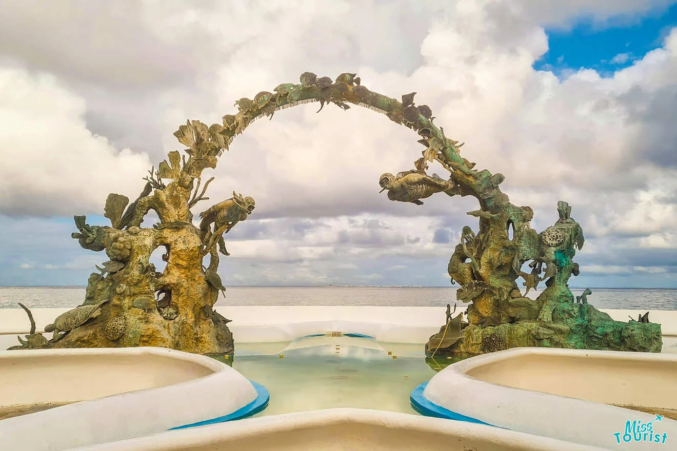 An ornate, marine-themed metal arch sculpture with various sea creatures stands against a cloudy sky, reflected in a shallow pool.