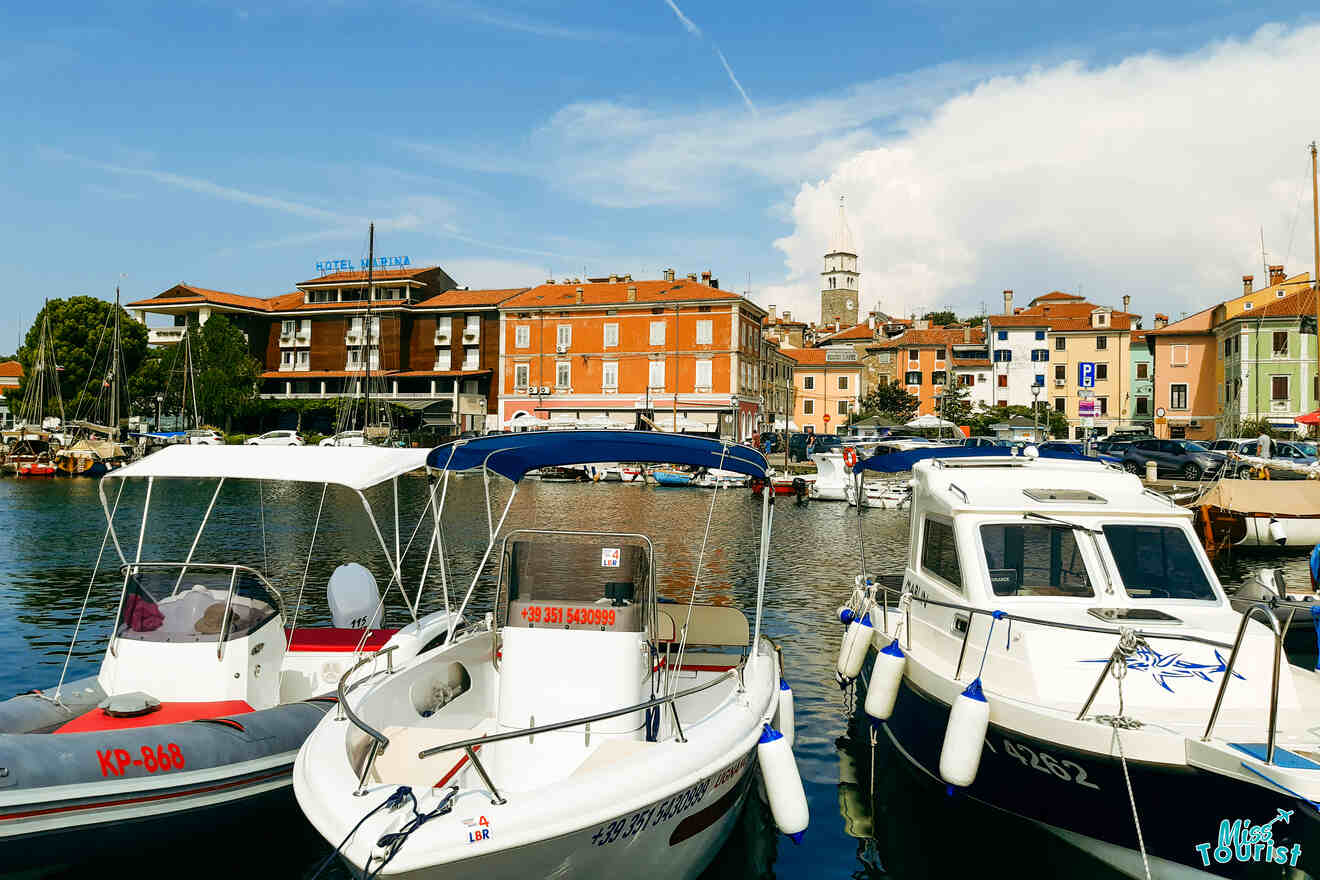 Small boats docked in a marina with a view of colorful buildings and a clock tower in the background under a partly cloudy sky.