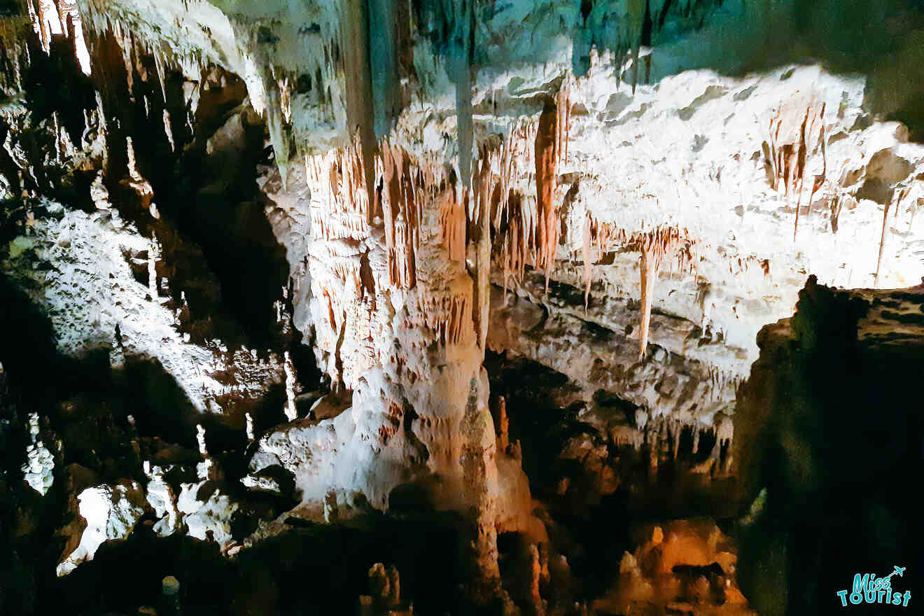 A dimly lit cave interior with numerous stalactites hanging from the ceiling and stalagmites rising from the ground. Shadows cast a dramatic effect on the rock formations.