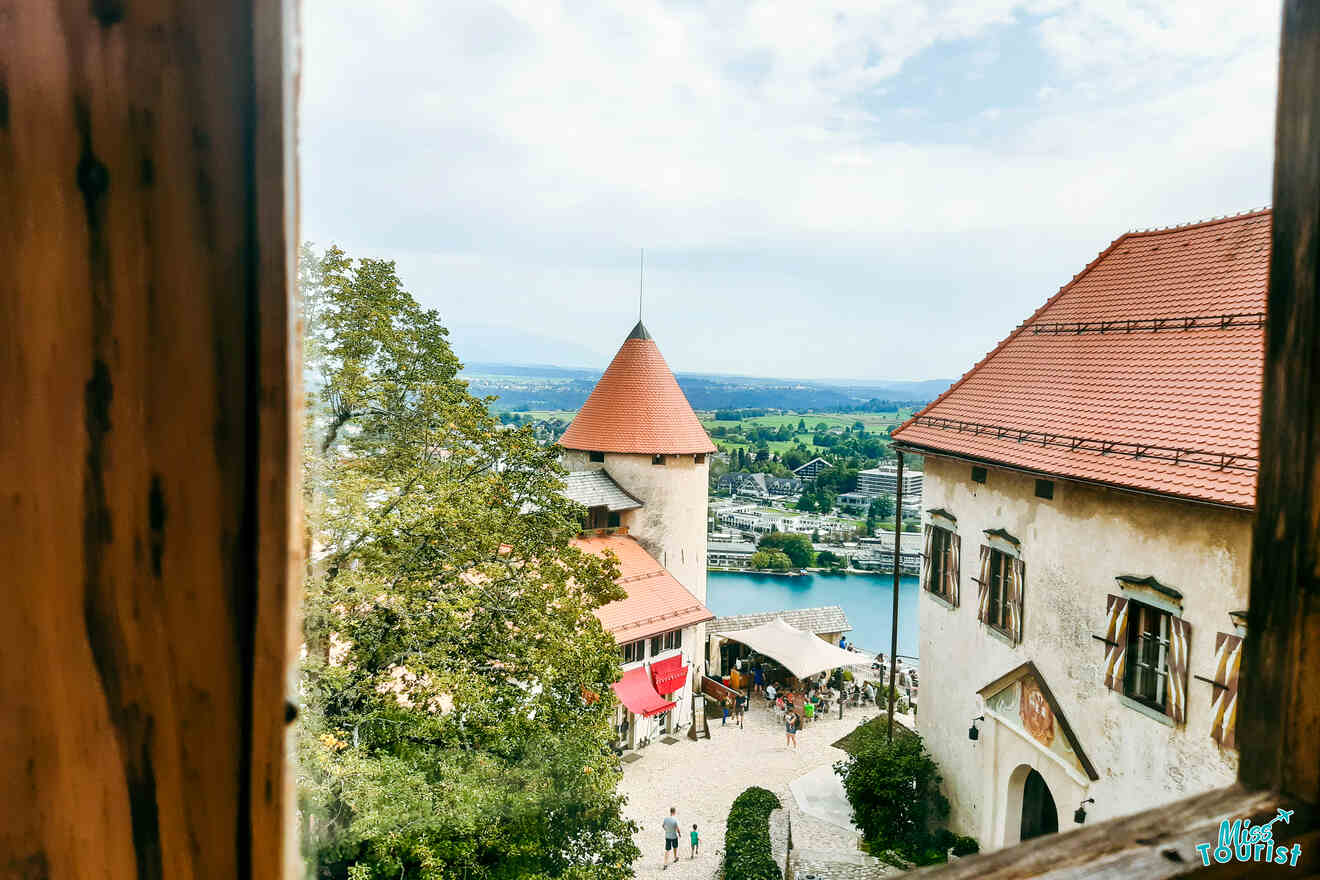 View through a window of a medieval castle courtyard with red-tiled roofs, a conical tower, and people gathered below, surrounded by trees and overlooking a distant scenic landscape.