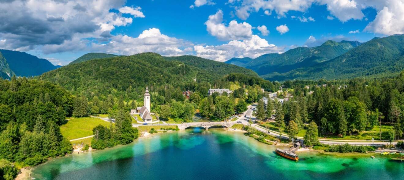 Aerial view of a vibrant lake surrounded by lush green hills and forests. A church with a tall steeple, a bridge, and a few buildings are visible near the water's edge under a partly cloudy sky.