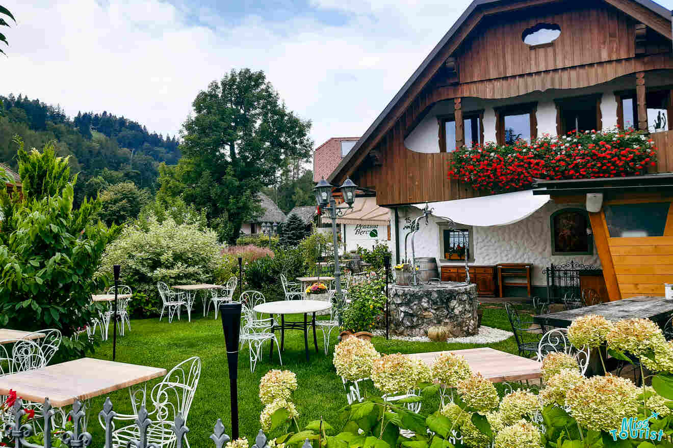 A rustic outdoor cafe with metal tables and chairs on grass, surrounded by greenery and flowers. A wooden building with red flowers on the balcony is in the background.