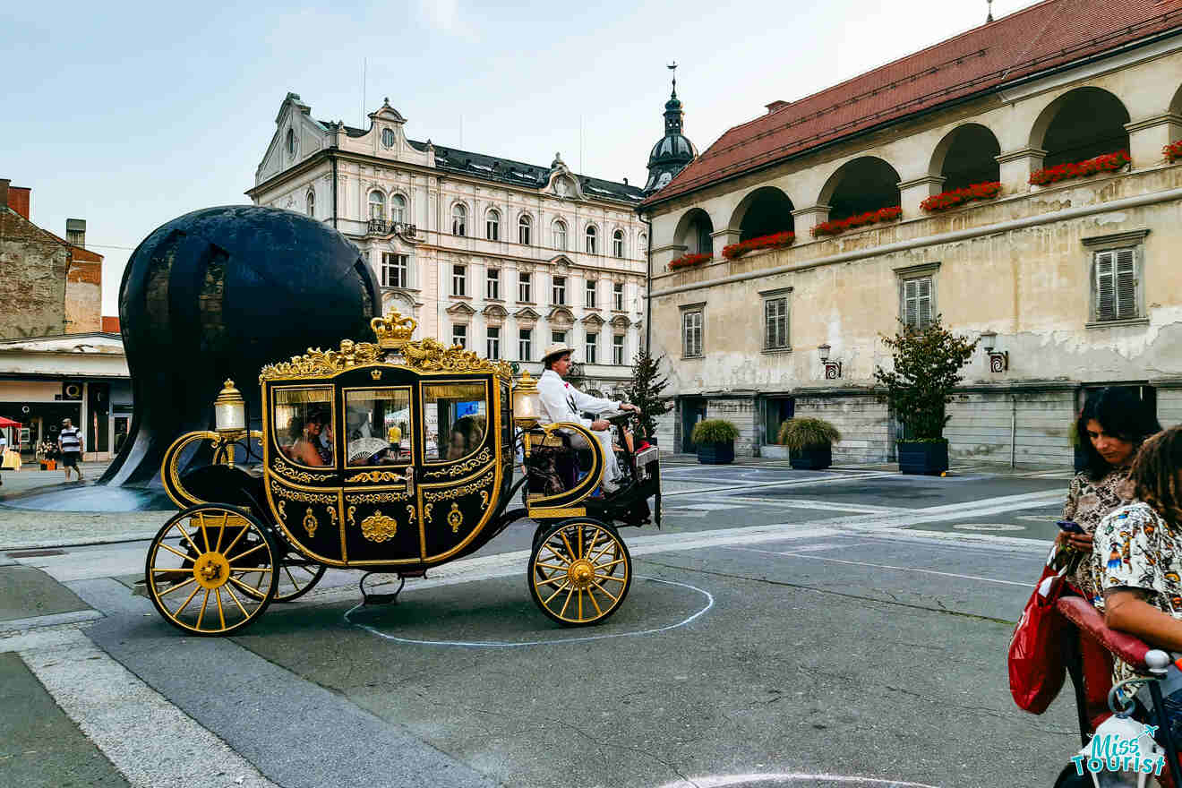 A decorative horse-drawn carriage passes through a European city square, surrounded by historic buildings and people walking.