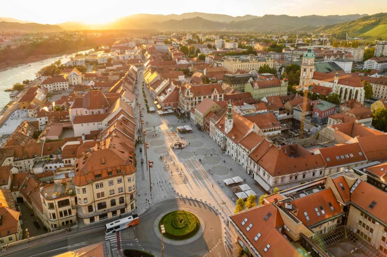 Aerial view of a European town with red-roofed buildings, a central square, and a river running along the left. The sun sets behind distant hills.