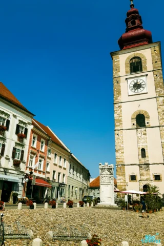 A cobblestone square with a large clock tower, surrounded by historic buildings. Flowers decorate balconies, and a bicycle is parked nearby. The sky is clear and blue.