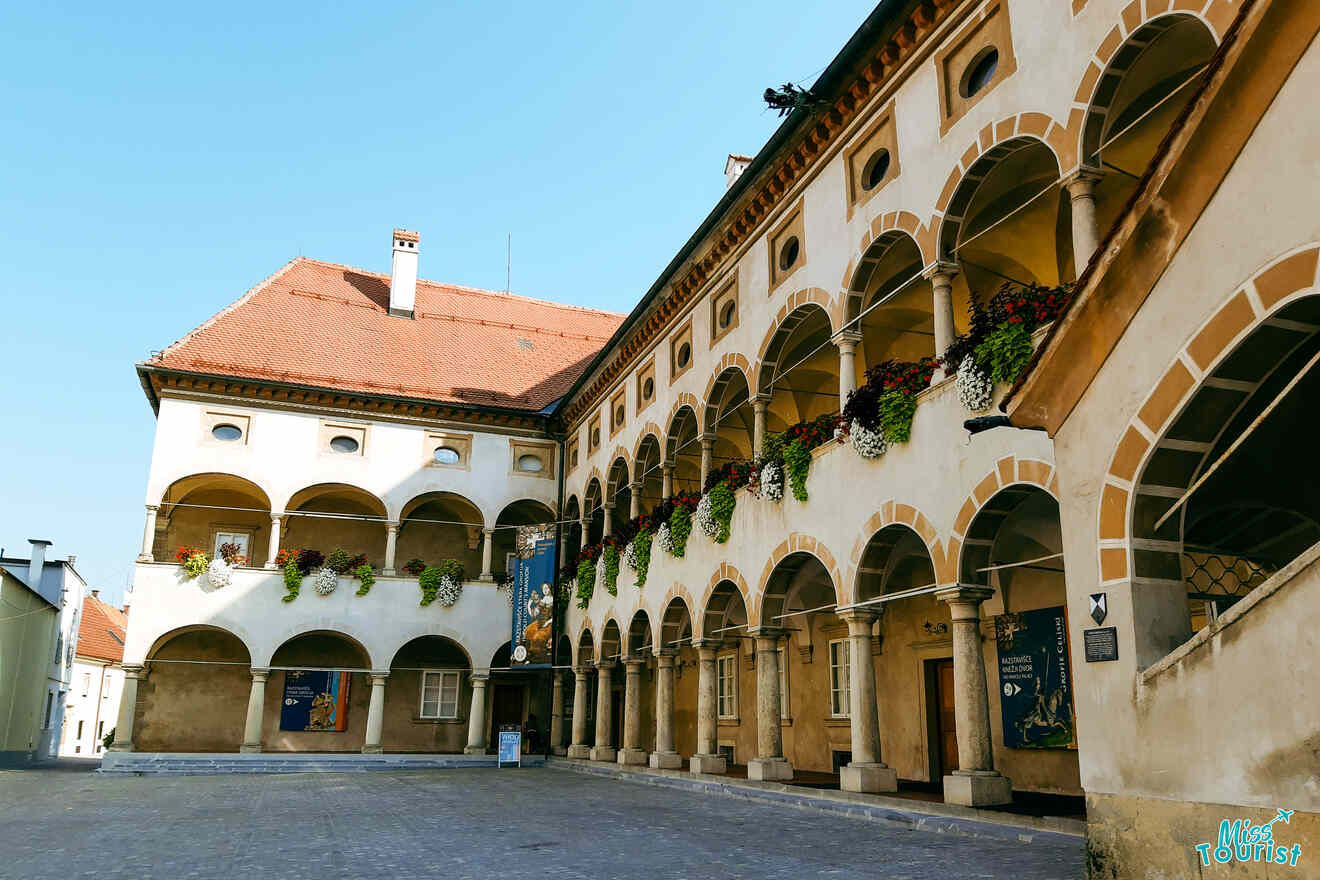 A historic building with arched walkways and a red-tiled roof. The facade features hanging flower pots and banners.