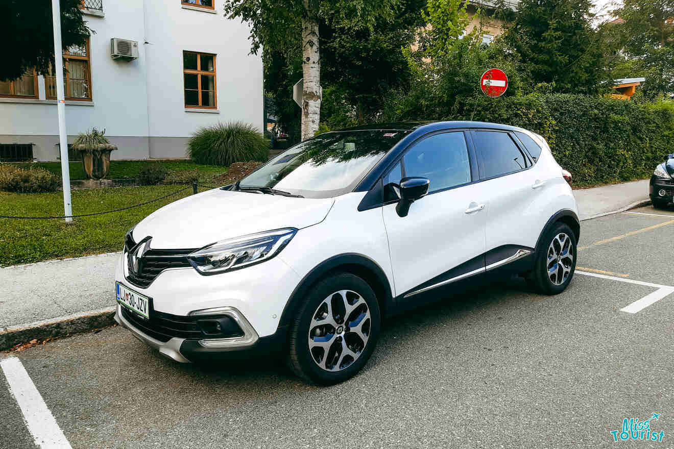 A white Renault Captur SUV is parked on the side of the street near a stop sign, with trees and buildings in the background.