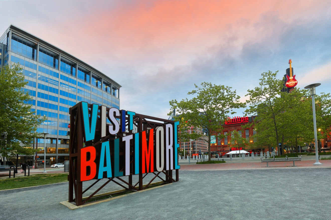 Large "Visit Baltimore" sign in front of modern buildings and trees, with a Hard Rock Cafe in the background under a cloudy sky.