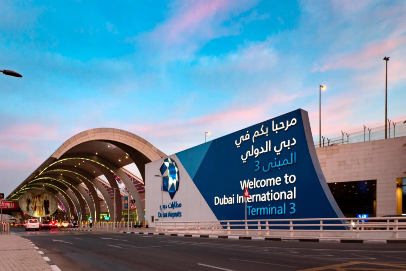 Dubai International Airport Terminal 3 entrance with welcome signage, illuminated under a colorful sky. Several taxis and vehicles are parked nearby.