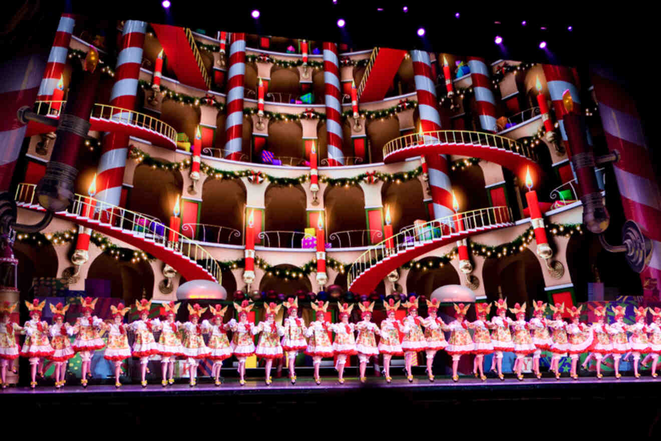 A large group of dancers in colorful costumes performs on stage with a vibrant, multi-level, holiday-themed backdrop featuring candy canes and festive decorations.