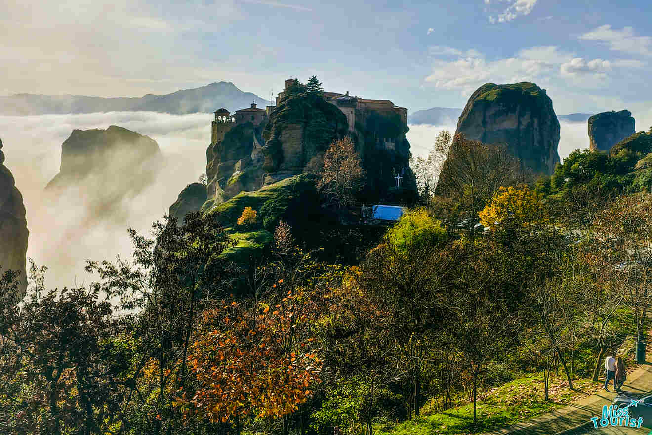 A monastery sits atop a towering rock formation surrounded by mist and lush vegetation, with mountains in the background under a partly cloudy sky. A person walks on a path in the foreground.