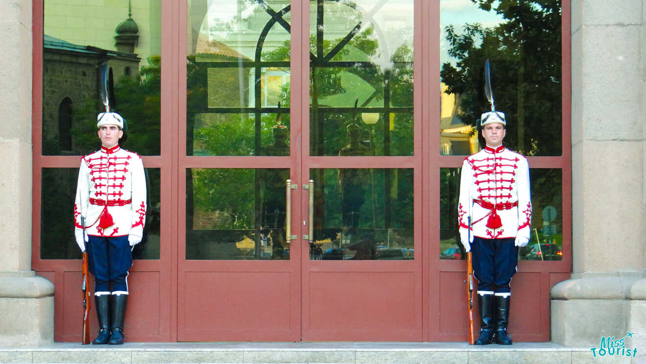 Two uniformed guards stand at attention outside a building with glass doors and a reflection of trees.