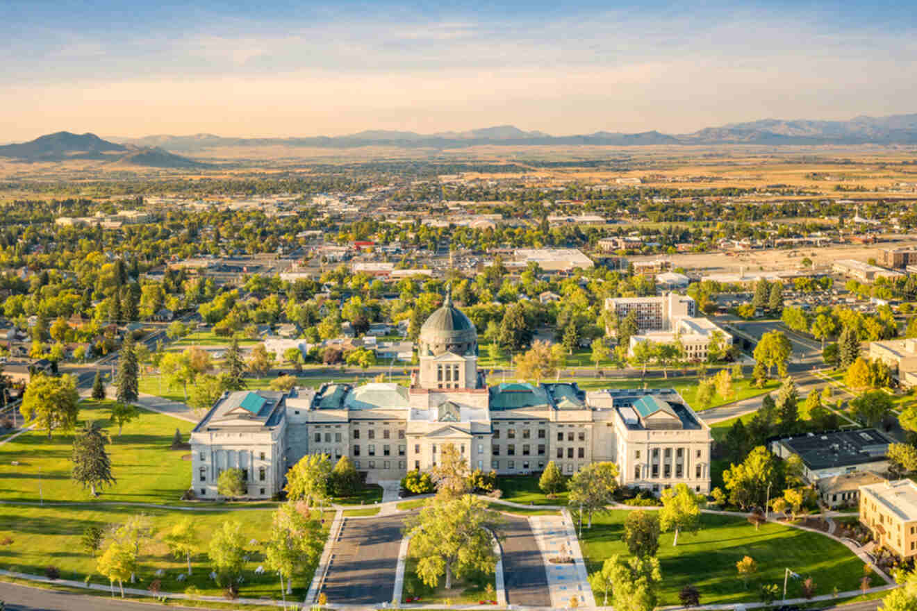 Aerial view of a large, historic building with a dome, surrounded by green lawns and trees, with a city and mountains in the background under a clear sky.