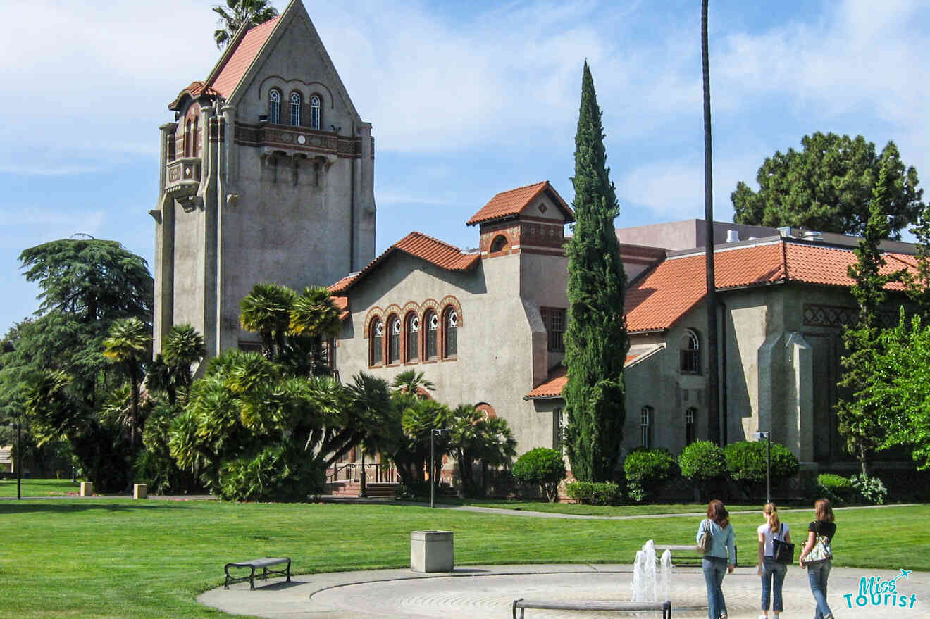 A historic building with a clock tower and red-tiled roof on a sunny day, surrounded by grass and trees. Three people are walking near a fountain in the foreground.