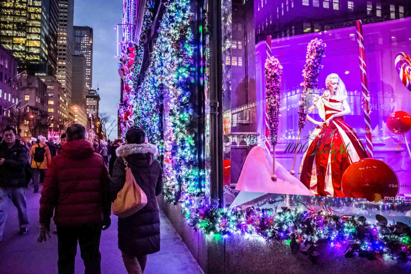 People walking past a brightly decorated holiday window display on a city street at dusk, with colorful lights and festive ornaments.