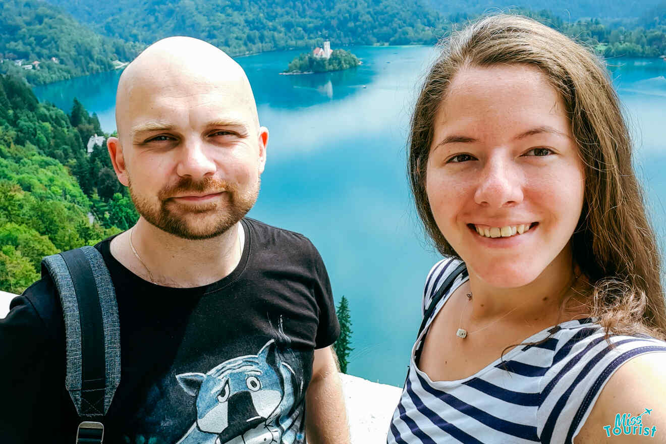 The writer of the post with her husband take a selfie in front of a scenic lake with a small island and a building on it. Green hills surround the lake.