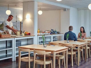 A man prepares food at a counter while a man and woman sit and talk at a table in a bright, modern dining area with wooden chairs and tables.