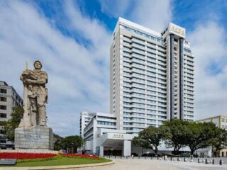A tall modern hotel building stands behind a large stone statue of a soldier with trees and blue sky in the background.