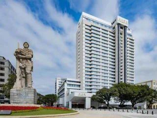 A tall modern hotel building stands behind a large stone statue of a soldier with trees and blue sky in the background.