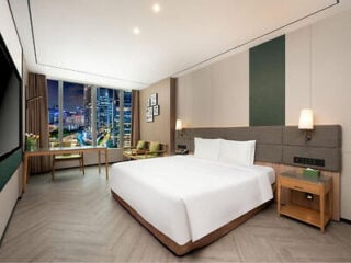 Modern hotel room with a large bed, nightstands, a desk, two chairs, and a large window showing a cityscape at night.