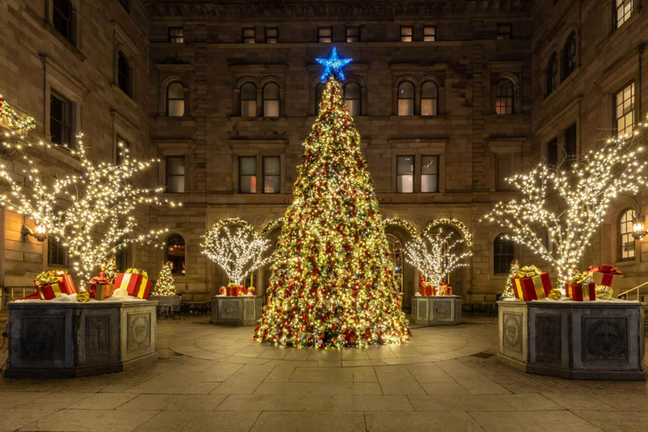 A large, decorated Christmas tree with glowing lights and a blue star topper is surrounded by lit trees and wrapped gifts in a courtyard.