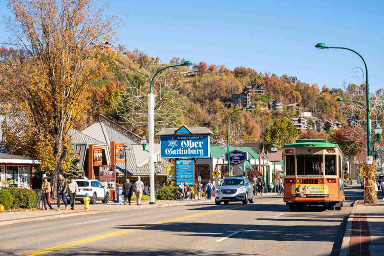 Street scene in Gatlinburg with a trolley, shops, and people walking. Trees and hills with autumn foliage are in the background.