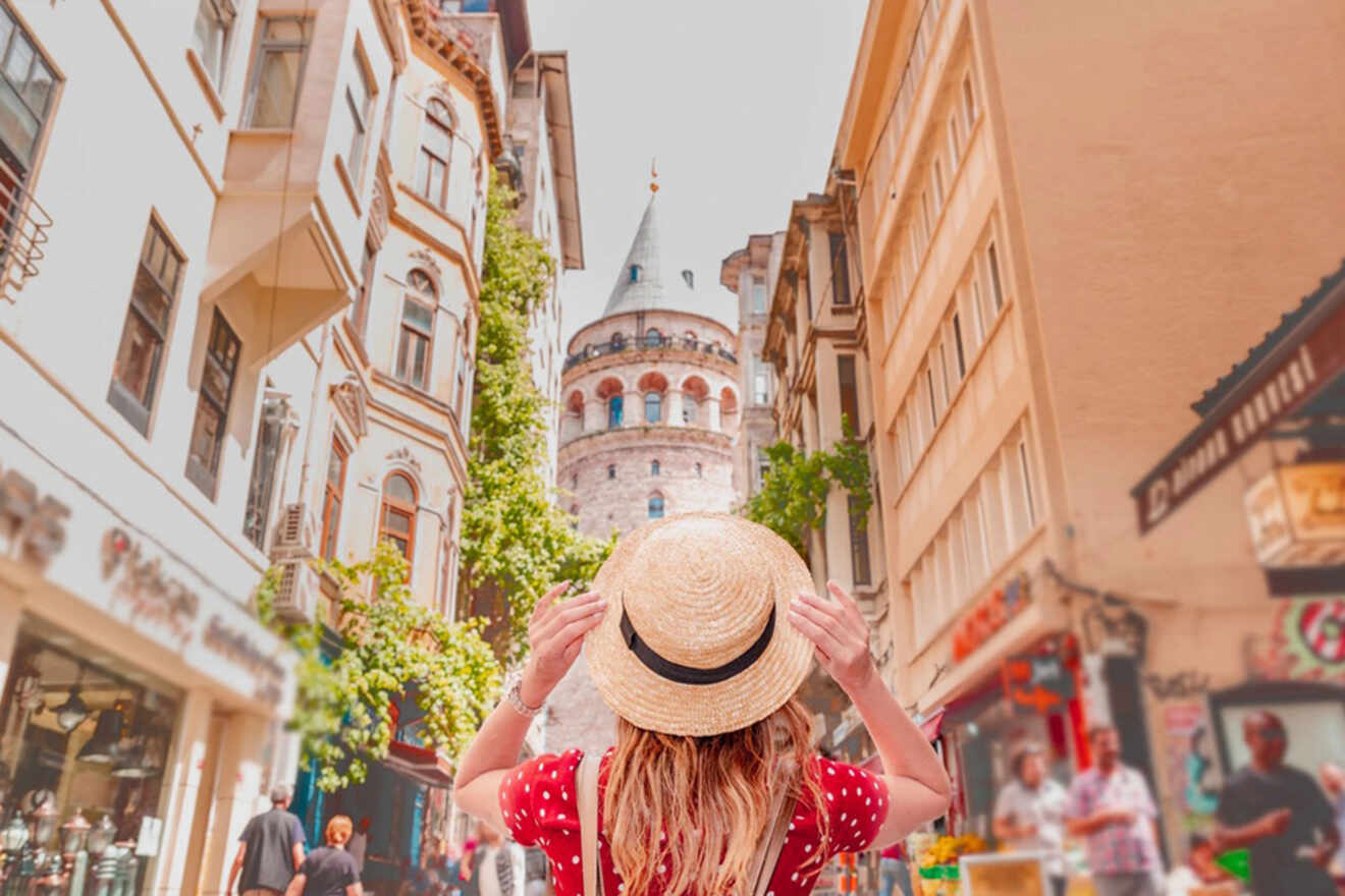 A person in a red polka dot dress holds a straw hat while looking at the Galata Tower on a sunny street with shops and pedestrians.