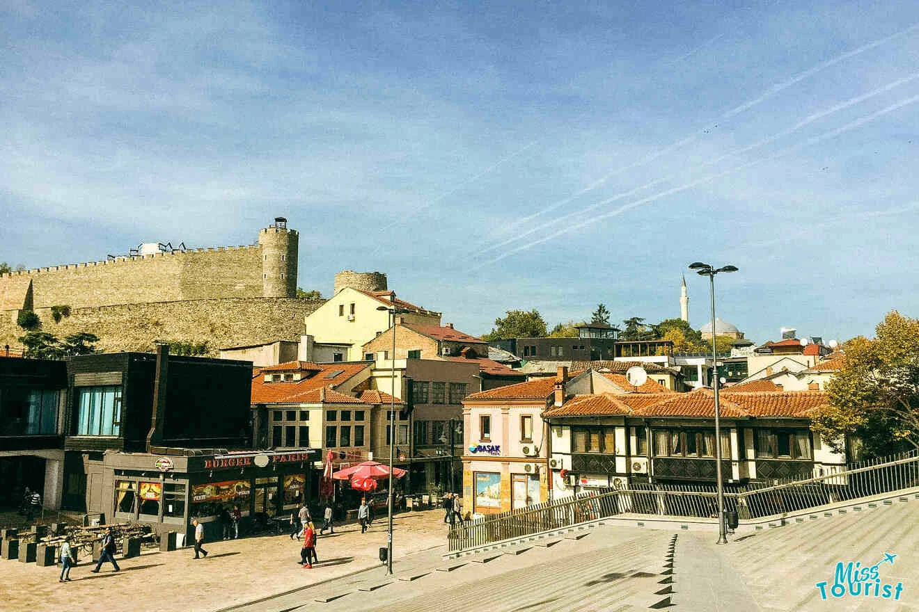 A view of a town square with a castle on a hill in the background, surrounded by buildings and a few people walking.