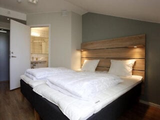 Neatly made double bed with white bedding and wooden headboard in a modern hotel room; open door reveals a bathroom with a sink and shower.