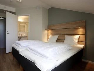 Neatly made double bed with white bedding and wooden headboard in a modern hotel room; open door reveals a bathroom with a sink and shower.