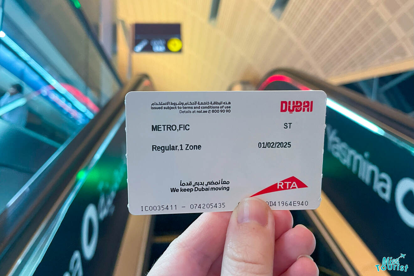 A hand holds a Dubai metro ticket in front of an escalator, displaying the date 01/02/2025 and marked for a Regular, 1 Zone journey.